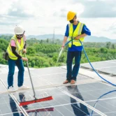 Two technicians wearing hard hats and safety vests cleaning rooftop solar panels with a water hose and a long brush.